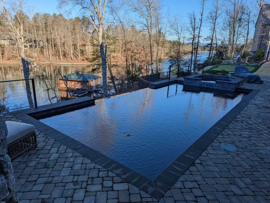 Dark infinity edge pool and raised spa on a paver deck, looking out over a lake