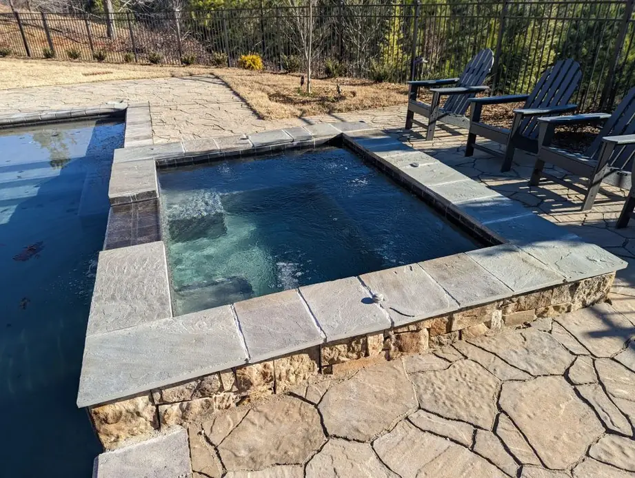 Square stone-coped spa beside a pool with Adirondack chairs on the deck